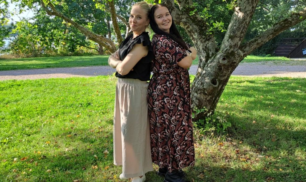 Kuvassa Kirkkopalvelujen varainhankinnan päällikkö Ruut Lemmetyinen ja Kotimaanavun koordinaattori Elisa Huuhka seisovat ulkona puun alla selät vastakkain, aurinko paistaa. 